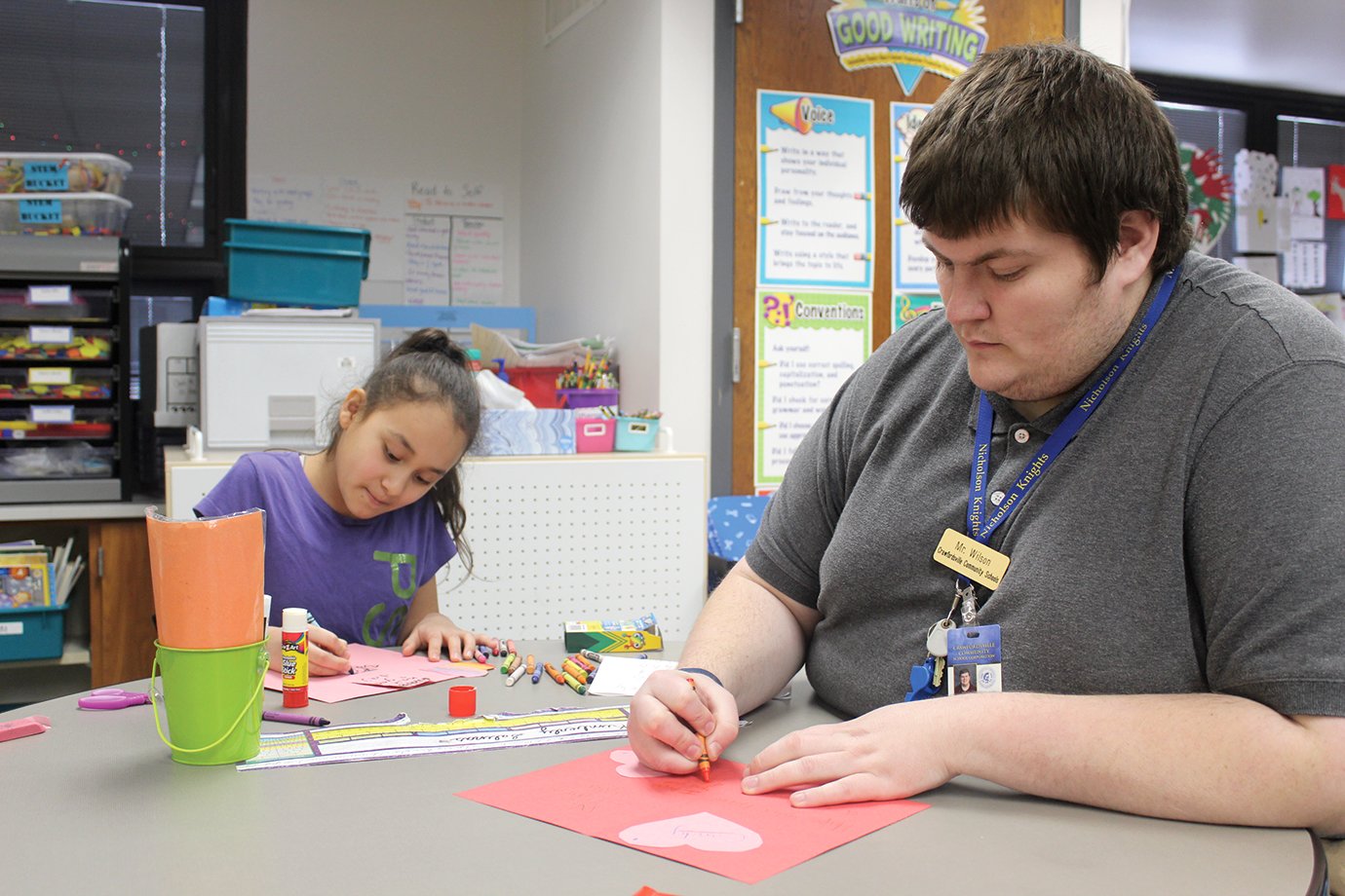 Tanner Wilson, third-grade teacher at Nicholson, sits with student Kimberly Salinas while creating Valentine”s Day cards on Thursday.