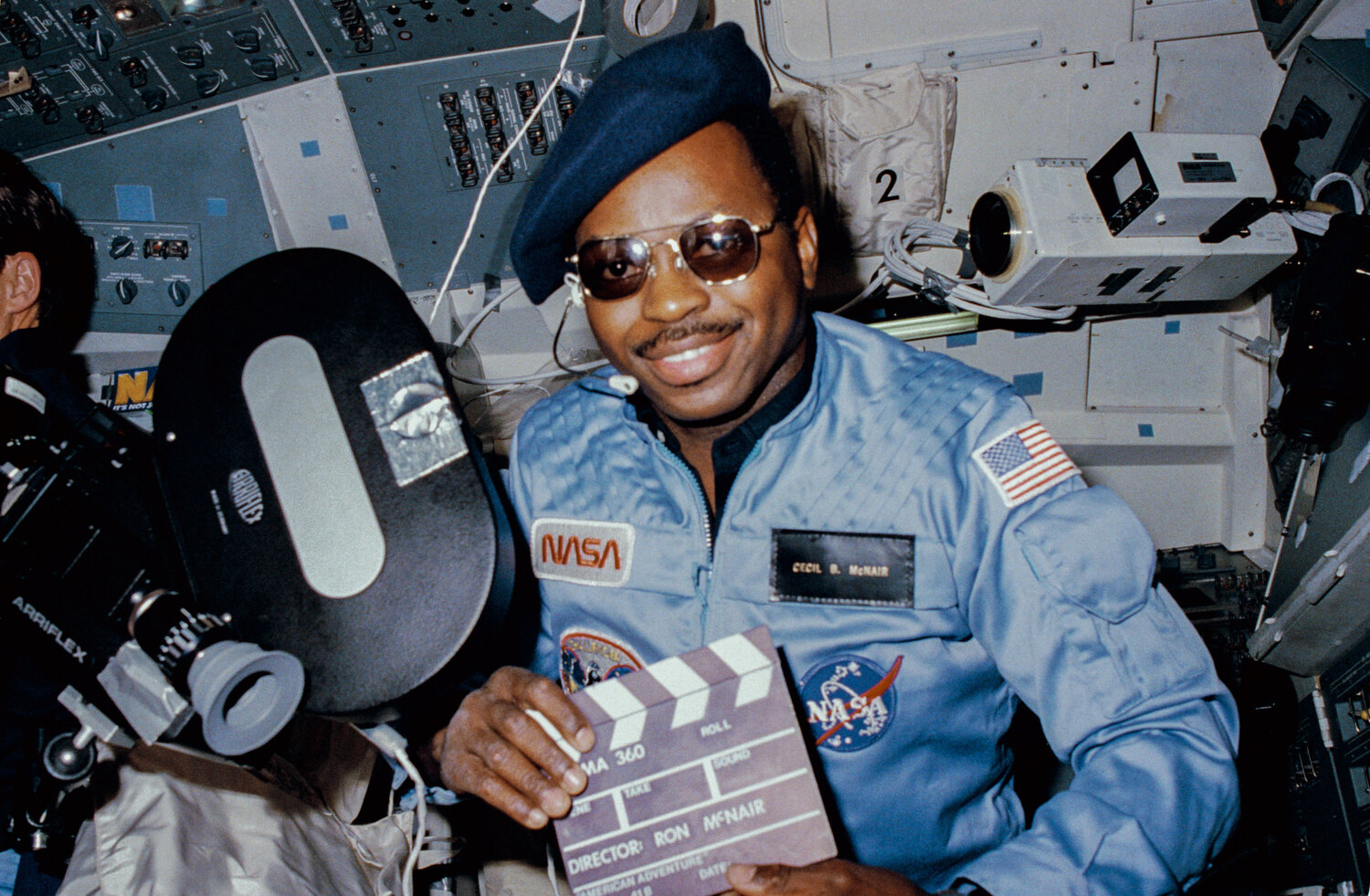 Astronaut Ronald E. McNair, 41-B mission specialist, doubles as "director" for a movie being "produced" aboard the Earth-orbiting Space Shuttle Challenger in February 1984. McNairís name tag ("Cecil B. McNair"), beret and slate are all humorous props for application of a serious piece of cargo on this eight day flight - the Cinema 360 camera. Two of the cameras were carried aboard the Challenger to provide a test for motion picture photography in a unique format designed especially for planetarium viewing. This camera was located in the crew cabin area and a second was stowed in a getaway special (GAS) canister in the payload bay.