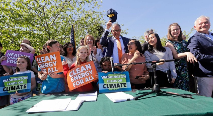 Washington Gov. Jay Inslee, center, pulls off his “100%” cap, standing for a goal of 100% clean electricity, after posing for a photo with supporters after signing climate protection legislation on May 7, 2019, in Seattle. As a first step toward getting fossil fuels out of electricity, the measure required utilities to stop burning coal by the end of 2025.