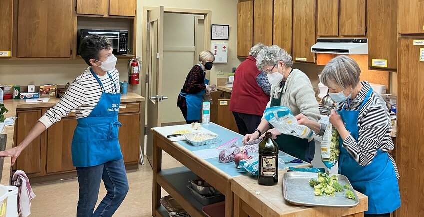 Sally Brennand, Lee Doyle, name omitted, Sally Alhadeff, and Anne Radford, preparing food and wearing their signature blue aprons, "OUUC Feeds People."