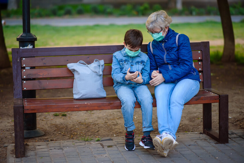 Grandmother with grandson outdoors in park in medical masks sit on a bench with smartphone. Boy shows something on screen. Chatting or playing game. Quarantine covid-19, pandemic coronavirus 2020