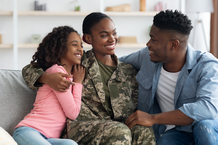 A black woman veteran in fatigues with her teen daughter and husband having a happy conversation sitting on a couch.