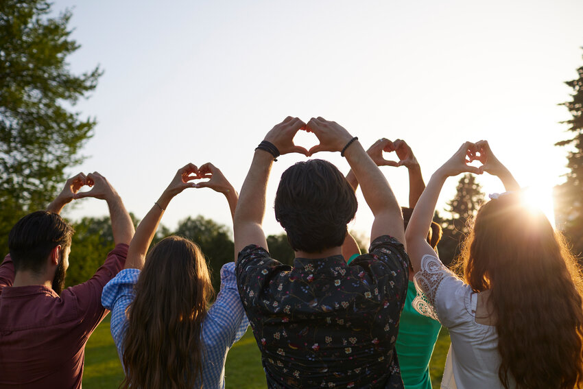 A group of people holds hands with a heart sign on a sky background with sunset in a park.