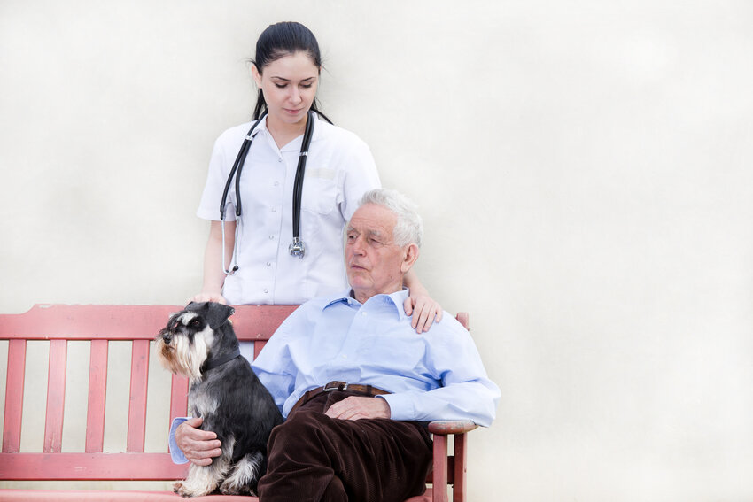Senior man with dog sitting on the bench while younger woman doctor standing behind him and holding her hand on his shoulder