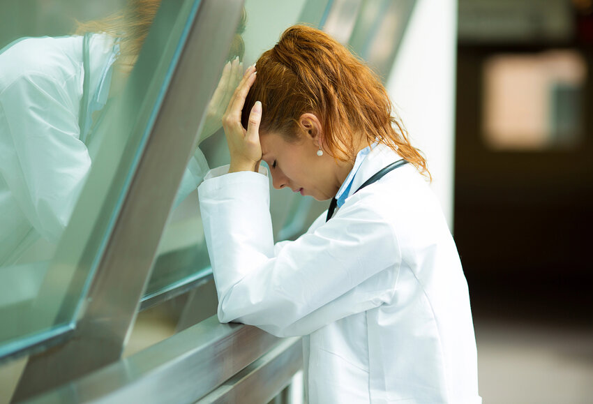 An exhausted woman doctor leaning up against the glass, holding her head.