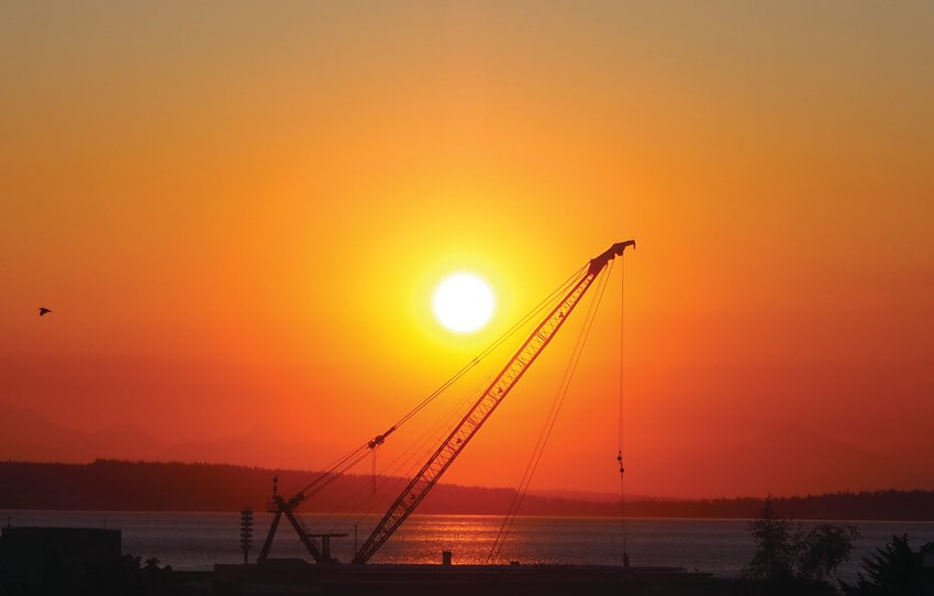 The derrick crane at the ongoing breakwater project at Point Hudson is silhouetted by the rising sun. Port officials say the work is proceeding at a dependable pace.