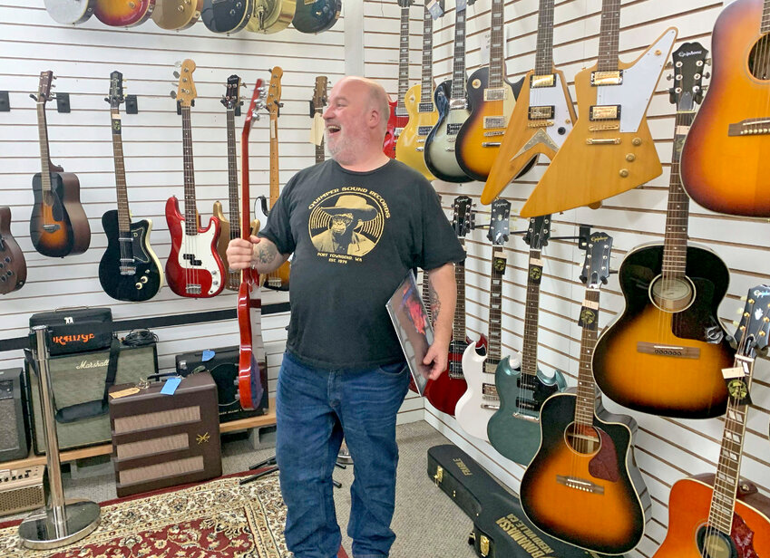 Quimper Sound owner James Schultz stands in front of store&rsquo;s guitar wall, holding a Black Sabbath album and a Gibson SG endorsed by the band&rsquo;s guitarist, Tony Iommi.