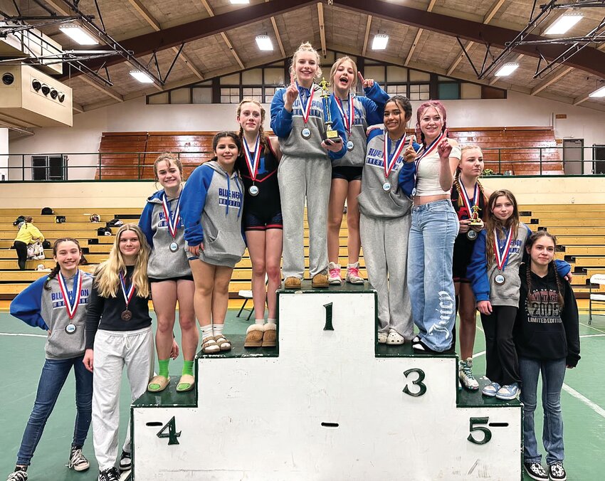 A proud group of girls stands at the podium showing their medals and team championship trophy. From left: Angie Luckhurst, Brooke Seton, Ella Welte, Natalia “Natalie” Duran, Katalena Perka, Leighton Dunn, Layla Grimm, Elizabeth “Liz” Camacho, Riley Eubanks, Kindle Iverson, Ayla Brayton and Ruby Lehman. Not in the photo but competing at the event were Elizabeth “Eli” Duran Paniagua, Ariana Vergara and Violet Weidner.  The Rivals girls wrestling squad has now won this season-ending tournament two years in a row.
