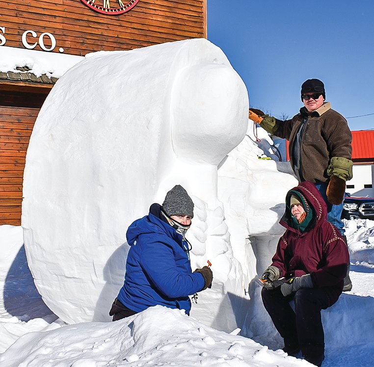 Jaymie Stocks (left), Sarah Moldenhauer (center), and Mason Wiekert 
(standing) carved the 
traditional snow sculpture on Monday depicting this year’s pin for the Ely Winter Festival.