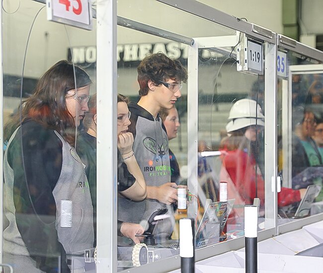 The Iron Mosquitos drive team looks on as Greyson Reichensperger controls the team’s robot from behind the plexiglass panels of the control booth.
