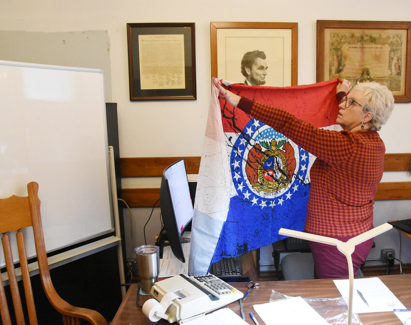 Gasconade County Clerk Lesa Lietzow opens up a Missouri flag Dec. 28 she was planning on using to cover an unpainted wall behind the screen (left) where a book shelf once occupied a corner of the Commission meeting room. The screen separates her new desk location with an area planned for in-person absentee voting. Her move into the meeting room was necessitated by plans to use a storage room for an elevator being built inside the courthouse and the need for additional staff office space in the County Clerk&rsquo;s office.