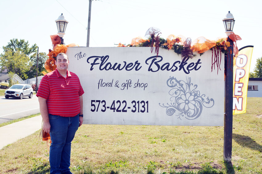 The Flower Basket’s new owner Cole Higbie poses beside the sign outside the business.