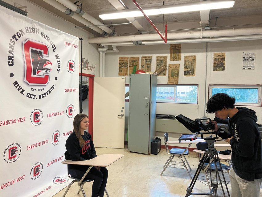 ON AIR: Eric Zhang readies the camera while Gisele Rotolu sits in front of Cranston West&rsquo;s backdrop gearing up to read from the teleprompter. (Herald photo)