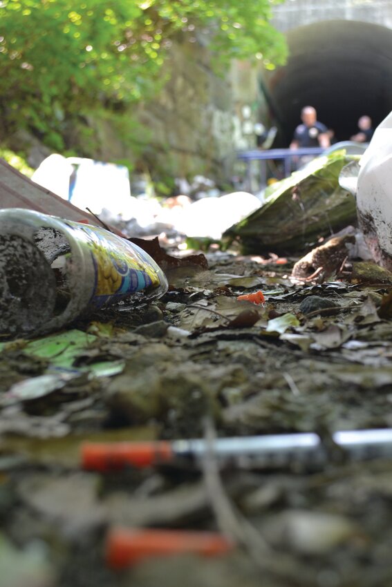 NEEDLE CARPET: In the background, Cranston Police Capt. Justin Dutra and Special Victims Unit Detective Mike Iacone can be seen visiting a makeshift camp in a tunnel behind Cranston Police Headquarters. In the foreground, one of hundreds of needles littering the campsite.