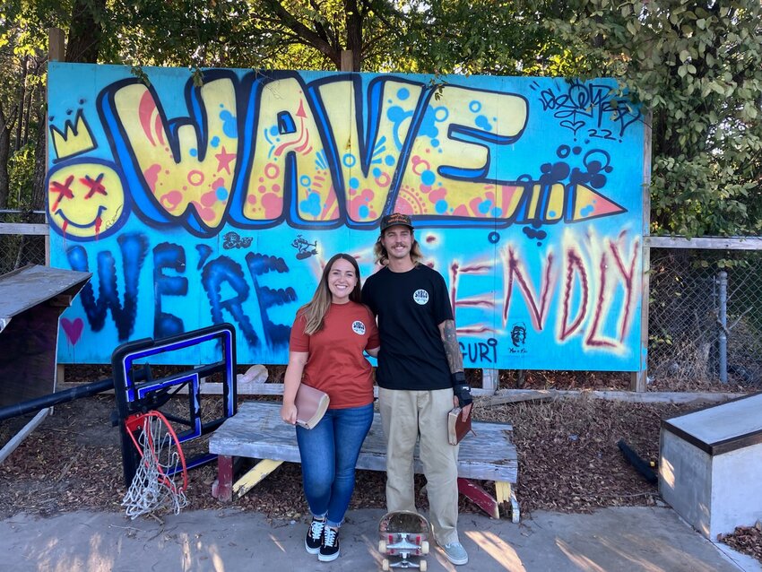 Austin and Haeley Arnold in front of the skate park&rsquo;s &ldquo;Wave, we&rsquo;re friendly&rdquo; sign with their Bibles and Austin&rsquo;s skateboard.